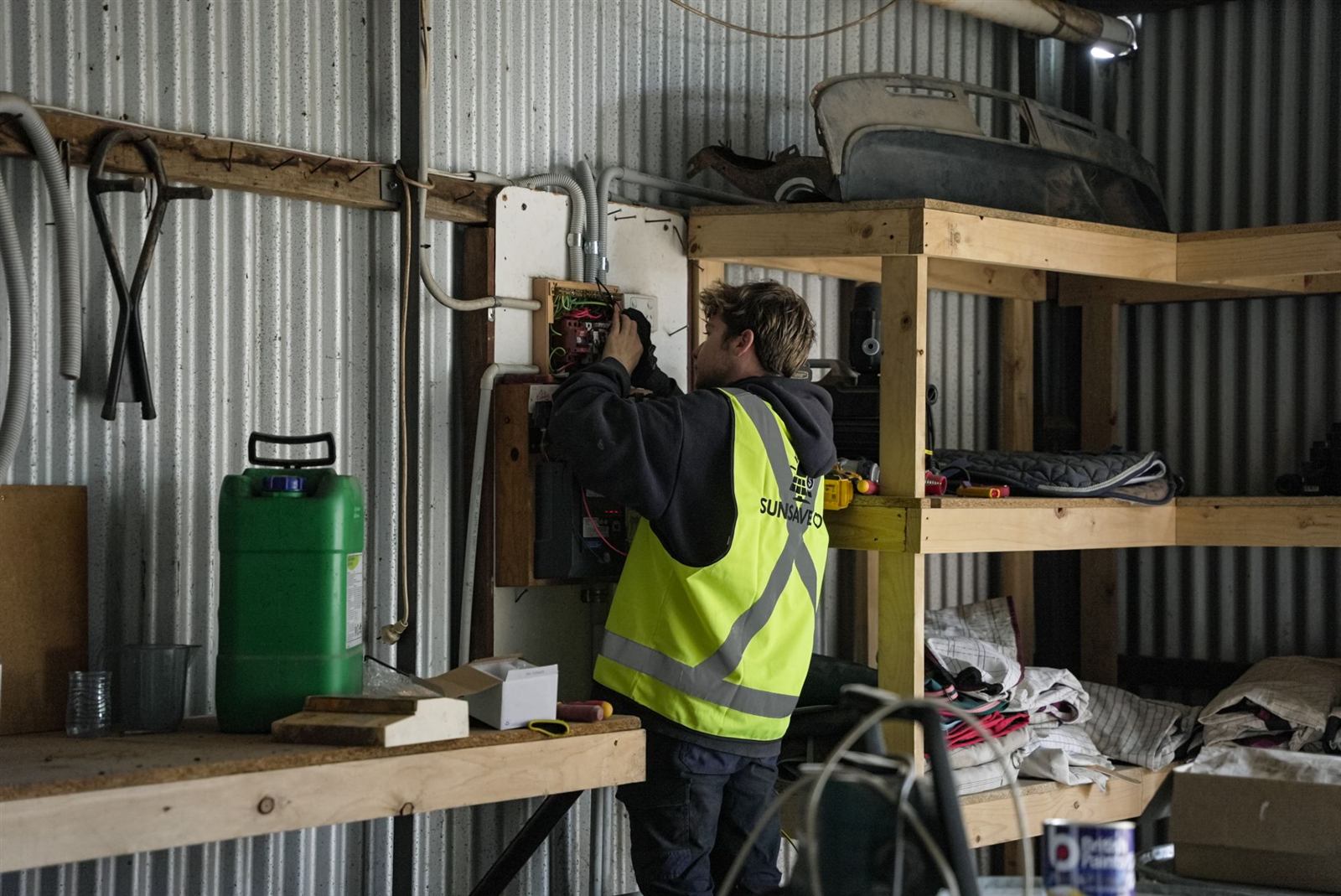 A Sunsave electrician in a hi-vis vest working on a switchboard inside a corrugated-iron shed
